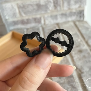 May include: Two black plastic cookie cutters, one shaped like a star and the other shaped like a circle with a scalloped edge.