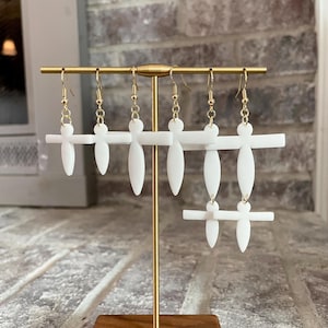 May include: White cross-shaped earrings with gold hooks hanging on a gold stand. The earrings are in a minimalist style.