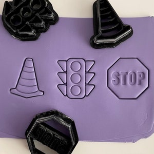 May include: Purple silicone baking mat with five black plastic cookie cutters. The cutters are shaped like a traffic light, a stop sign, two traffic cones, and a hexagon.