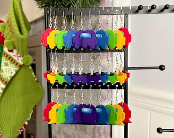 May include: A display of colorful Among Us character earrings. The earrings are made of polymer clay and feature different colors, including red, yellow, green, blue, and purple. The earrings are hanging on a black earring display stand.