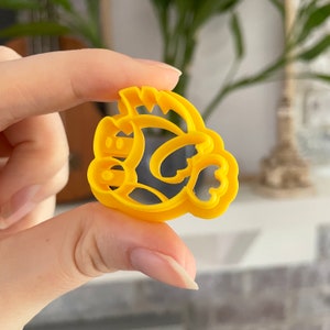 May include: A yellow plastic cookie cutter shaped like a cartoon character with a large head and a small body. The character has a large, round head with two small eyes and a wide, open mouth. The body is small and has two short arms and two short legs.