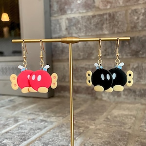May include: A pair of dangle earrings featuring a red and black cartoon character. The red character has white eyes and a yellow bow. The black character has white eyes and a yellow bow. The earrings are hanging on a gold stand.