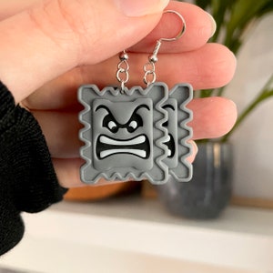 May include: A pair of gray earrings with a pixelated angry face design. The earrings are made of polymer clay and have silver hooks.