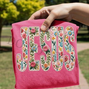 May include: A pink t-shirt with the word "JESUS" embroidered in colorful flowers. The letters are outlined with small white beads. The shirt is held up by a hand, with a blurred green background.
