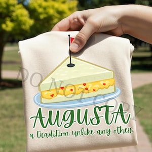 May include: A beige t-shirt featuring a cartoon sandwich on a blue plate with a toothpick and a red flag. The word "AUGUSTA" is written in green script, with the phrase "a tradition unlike any other" below.