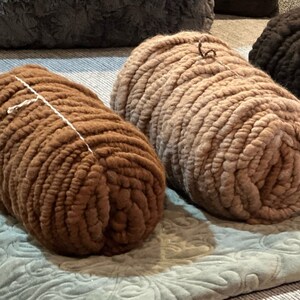 May include: Four rolls of yarn in different colors, including brown, beige, and dark brown. The yarn is arranged on a patterned blanket.