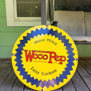 May include: A round metal sign featuring a yellow background with a dark blue jagged border. The text reads More Miles, Woco Pep, King of Motor Fuel, and Less Carbon in red and black lettering. The sign has small mounting holes on the edges.