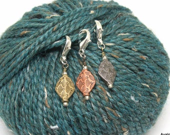 May include: Three metal yarn bowl stitch markers with a decorative diamond-shaped charm. The charms are in gold, copper, and silver with a wave pattern.