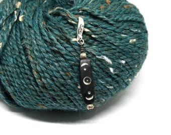 May include: A black and silver stitch marker with a moon and star design. The marker is hanging from a ball of teal yarn with white flecks.