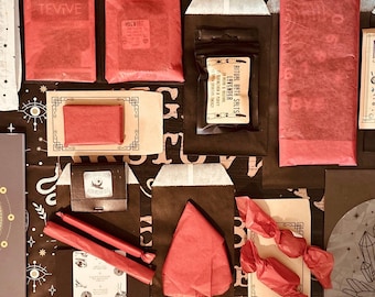 May include: An assortment of spiritual and self-care items arranged on a dark surface. Items include a journal, a bowl, wrapped packages, a small box, candles, and a book. The items are in various colors, including red, white, black, and gold.
