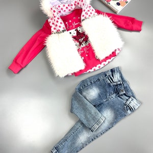 May include: A pink children's outfit featuring a hooded vest with white faux fur trim and red star pattern, a matching long-sleeved shirt with a "New York" graphic, and light-wash denim jeans. A floral notebook is also in the image.