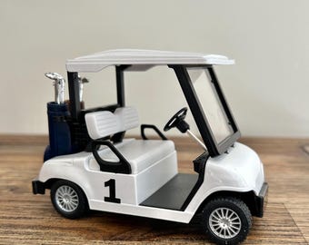 May include: A white miniature golf cart model featuring a black steering wheel, a white bench seat, and a dark blue golf bag containing silver golf clubs. The side of the vehicle displays the number 1 in black, and it has silver-colored wheels.