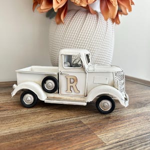 Peut inclure: Un camion en m&eacute;tal blanc de style vintage avec des roues noires et la lettre "R" sur la porte. Le camion est un objet d&eacute;coratif, avec une calandre avant d&eacute;taill&eacute;e et une petite benne. Un vase blanc avec des fleurs orange est en arri&egrave;re-plan.