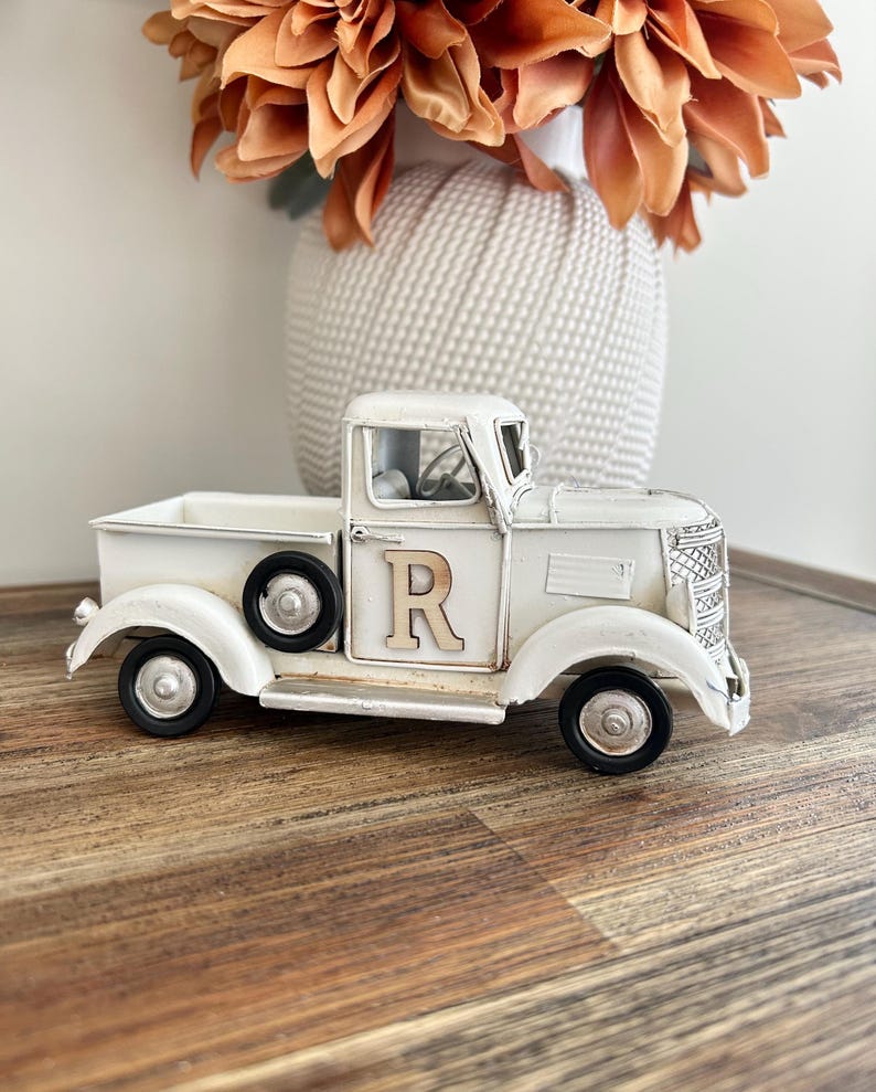 Peut inclure: Un camion en m&eacute;tal blanc de style vintage avec des roues noires et la lettre "R" sur la porte. Le camion est un objet d&eacute;coratif, avec une calandre avant d&eacute;taill&eacute;e et une petite benne. Un vase blanc avec des fleurs orange est en arri&egrave;re-plan.