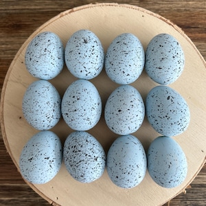 May include: Twelve light blue speckled decorative eggs arranged on a round wooden slice. The eggs have a speckled pattern in a darker shade of blue. The wooden slice has a natural wood grain.