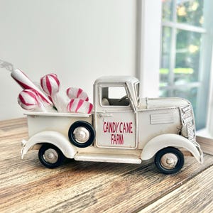 Könnte beinhalten: Ein weißer Spielzeug-LKW aus Metall mit schwarzen Rädern und dem in Rot auf die Tür gedruckten Text "CANDY CANE FARM". Die Ladefläche ist mit mehreren weiß-rot gestreiften, zuckerstangenförmigen Dekorationen in durchsichtiger Plastikverpackung gefüllt.