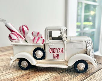 May include: A white metal toy truck with black wheels and the text "CANDY CANE FARM" printed in red on the door. The truck bed is filled with several white and red striped candy cane-shaped decorations wrapped in clear plastic packaging.