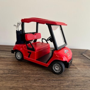 May include: A red miniature golf cart featuring a black roof, a clear windshield, and a black number 7 printed on the side. The vehicle includes a dark blue golf bag containing silver golf clubs at the rear and has black seats and silver wheels.