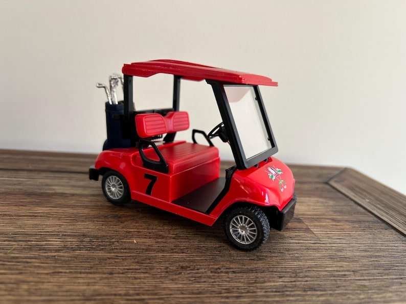 May include: A red miniature golf cart featuring a black roof, a clear windshield, and a black number 7 printed on the side. The vehicle includes a dark blue golf bag containing silver golf clubs at the rear and has black seats and silver wheels.