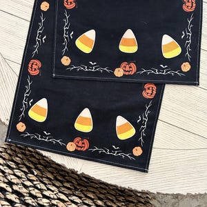 May include: A black table runner featuring a repeating pattern of orange jack-o'-lanterns, small orange pumpkins, white and yellow candy corn, and white silhouettes of bare tree branches and flying bats printed along the edges and center.