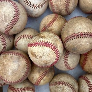 May include: A collection of well-worn baseballs. The baseballs are off-white with red stitching, showing signs of use. The image is a close-up, highlighting the texture and wear of the baseballs, suggesting they have been used in multiple games.