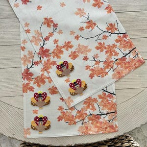 Puede incluir: Un camino de mesa de color crema con un patrón repetitivo de hojas de arce otoñales naranjas y rojas sobre finas ramas negras. Cuatro apliques bordados de pavos están sujetos a la tela, cada uno con un sombrero negro y un lazo de lunares rojos.