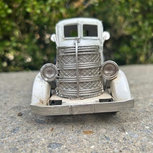 A small, white metal car model is displayed on a concrete surface, with a blurred green background.