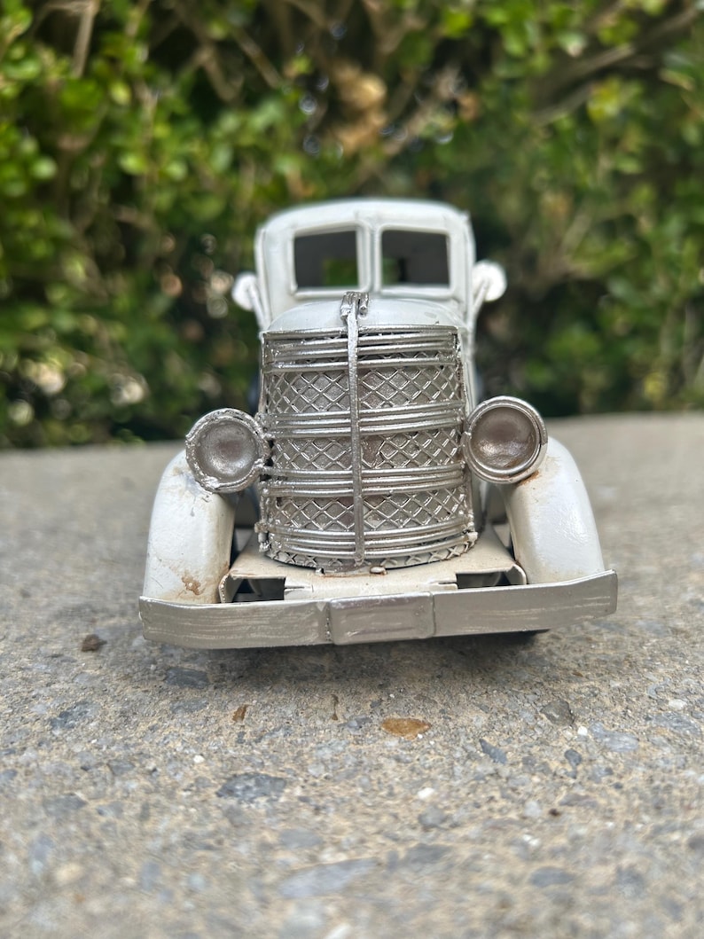 A small, white metal car model is displayed on a concrete surface, with a blurred green background.