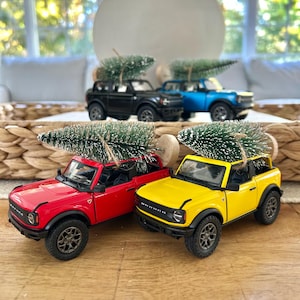 Puede incluir: Camiones de juguete en miniatura rojos, amarillos, negros y azules, cada uno con un pequeño árbol de Navidad en la parte superior. Los camiones están colocados sobre una superficie de madera, con un fondo neutro.