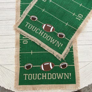 Puede incluir: Dos caminos de mesa de arpillera verde con diseño de campo de fútbol. Los caminos tienen líneas blancas, la palabra "TOUCHDOWN!" en blanco y adornos en forma de balón de fútbol. Los bordes están deshilachados con arpillera natural.