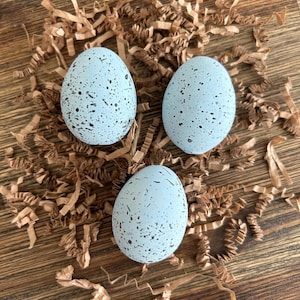 May include: Three speckled blue Easter eggs in a nest of brown shredded paper.