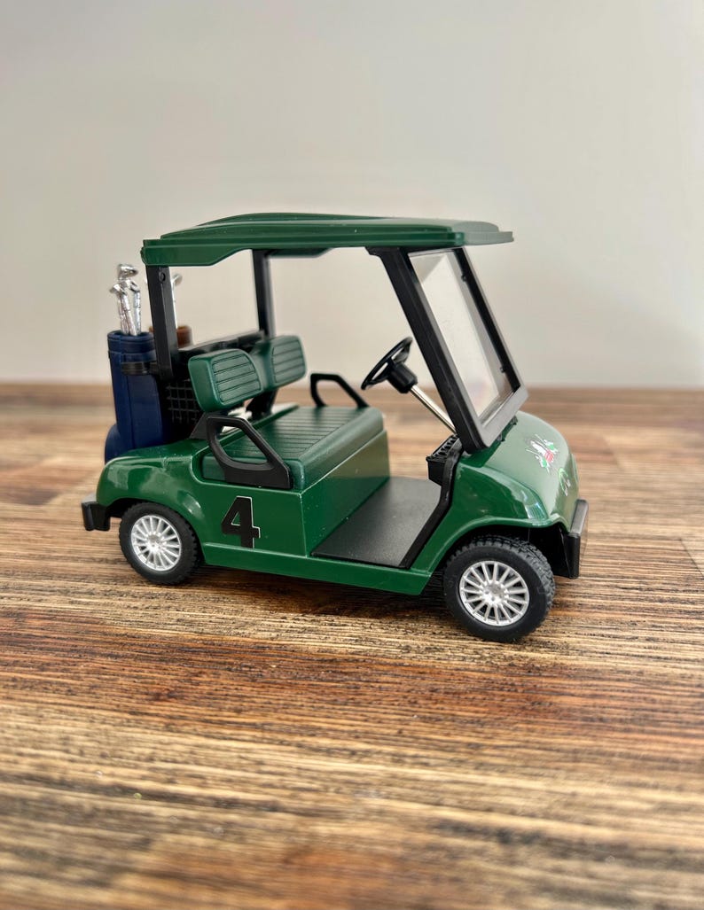May include: A miniature dark green golf cart model featuring a black steering wheel, a clear windshield, and a dark blue golf bag containing silver clubs attached to the rear. The side of the vehicle displays the number 4 in black lettering.