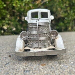 A small, white metal car model is displayed on a concrete surface, with a blurred green background.