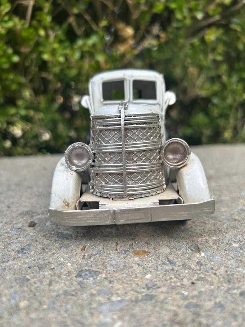 A small, white metal car model is displayed on a concrete surface, with a blurred green background.