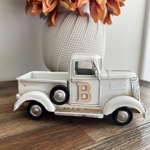 Peut inclure: Un camion vintage en m&eacute;tal blanc vieilli avec des roues noires et la lettre "B" en bois sur la porte. Le camion est pos&eacute; sur une surface en bois, avec un vase de fleurs orange en arri&egrave;re-plan.