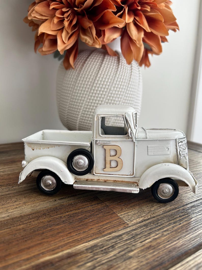 Peut inclure: Un camion vintage en m&eacute;tal blanc vieilli avec des roues noires et la lettre "B" en bois sur la porte. Le camion est pos&eacute; sur une surface en bois, avec un vase de fleurs orange en arri&egrave;re-plan.