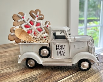 Op de afbeelding: Een witte metalen speelgoedtruck met zwarte wielen en de tekst BAKERY FARMS op de deur. De laadbak is gevuld met lichtgekleurd stro, een houten lepel en een grote bruine peperkoek-sneeuwvlokkoek versierd met wit en rood glazuur.