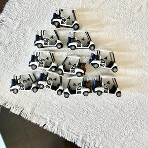May include: A collection of ten miniature white golf carts arranged in a triangular formation. Each cart features a dark blue golf bag at the rear, a black steering wheel, and the number 80 printed in black on the side of the white body.