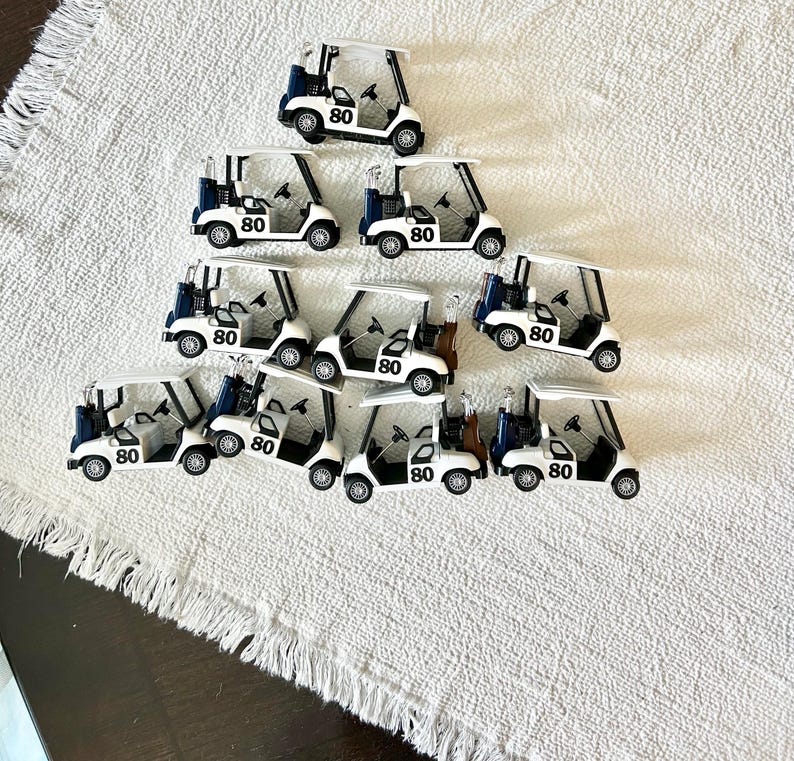May include: A collection of ten miniature white golf carts arranged in a triangular formation. Each cart features a dark blue golf bag at the rear, a black steering wheel, and the number 80 printed in black on the side of the white body.