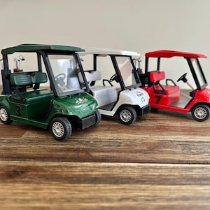 May include: Three miniature golf carts in dark green, white, and red. Each cart features a black roof, a steering wheel, a bench seat, and a rear rack holding miniature golf clubs. The green cart has the number 3, and the red cart has the number 7.
