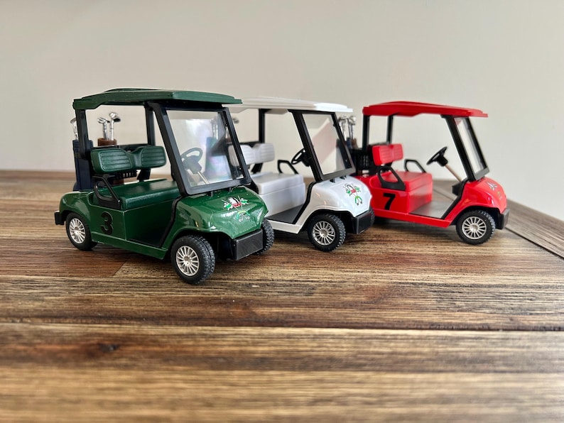 May include: Three miniature golf carts in dark green, white, and red. Each cart features a black roof, a steering wheel, a bench seat, and a rear rack holding miniature golf clubs. The green cart has the number 3, and the red cart has the number 7.