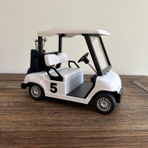 May include: A white miniature golf cart featuring a black number 5 on the side, a black steering wheel, a white bench seat, and a dark blue golf bag containing silver golf clubs positioned on the rear platform under the white roof structure.