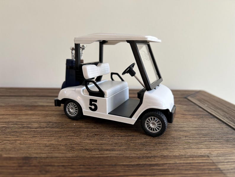 May include: A white miniature golf cart featuring a black number 5 on the side, a black steering wheel, a white bench seat, and a dark blue golf bag containing silver golf clubs positioned on the rear platform under the white roof structure.