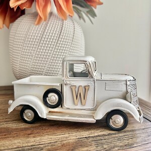 Peut inclure: Un camion en m&eacute;tal blanc de style vintage avec des roues noires et la lettre "W" en bois sur la porte. Le camion est un objet d&eacute;coratif, peut-&ecirc;tre un &eacute;l&eacute;ment de d&eacute;coration de style ferme. Un vase blanc avec des fleurs orange est en arri&egrave;re-plan.
