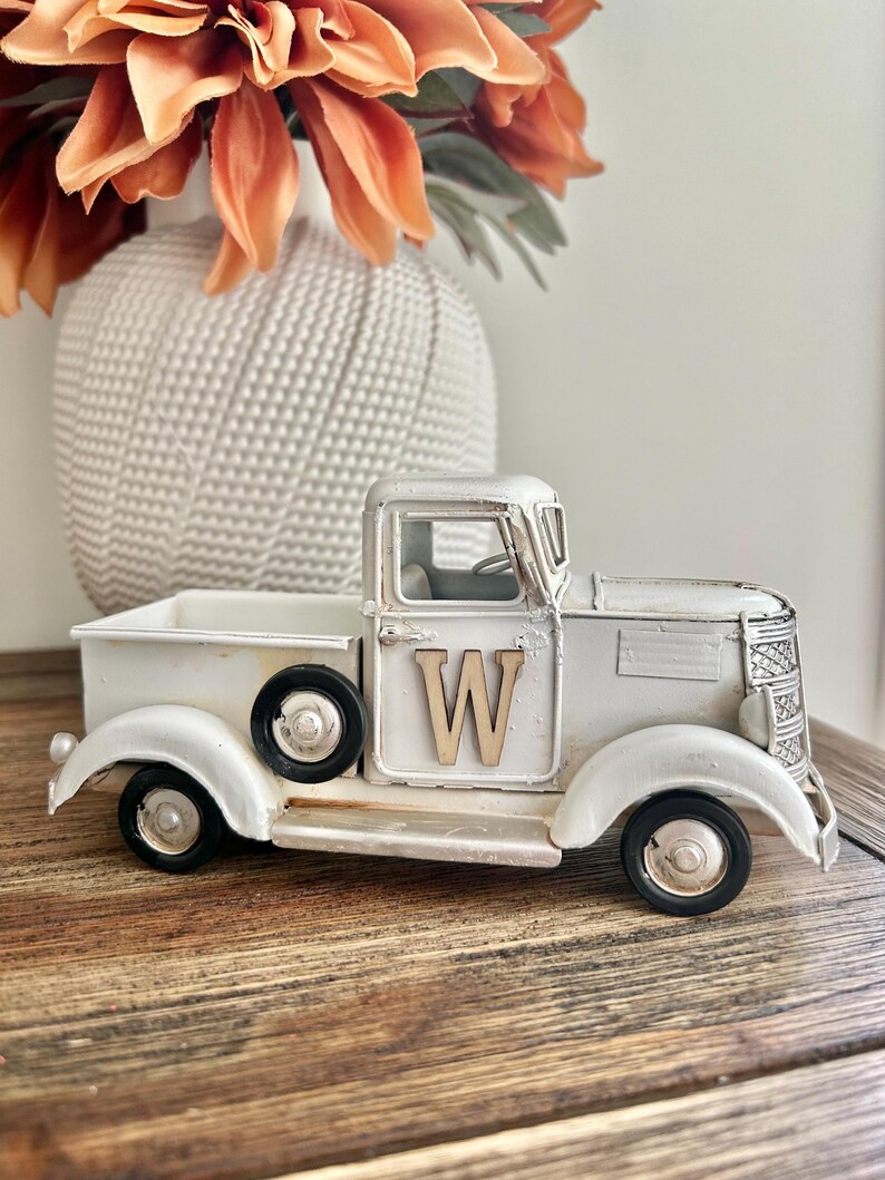 Peut inclure: Un camion en m&eacute;tal blanc de style vintage avec des roues noires et la lettre "W" en bois sur la porte. Le camion est un objet d&eacute;coratif, peut-&ecirc;tre un &eacute;l&eacute;ment de d&eacute;coration de style ferme. Un vase blanc avec des fleurs orange est en arri&egrave;re-plan.