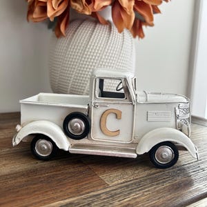 Peut inclure: Un camion en m&eacute;tal blanc de style vintage avec des roues noires et un "C" dor&eacute; sur la porte. Le camion est un objet d&eacute;coratif, peut-&ecirc;tre pour la d&eacute;coration int&eacute;rieure ou les expositions saisonni&egrave;res. Le camion est pos&eacute; sur une surface en bois.