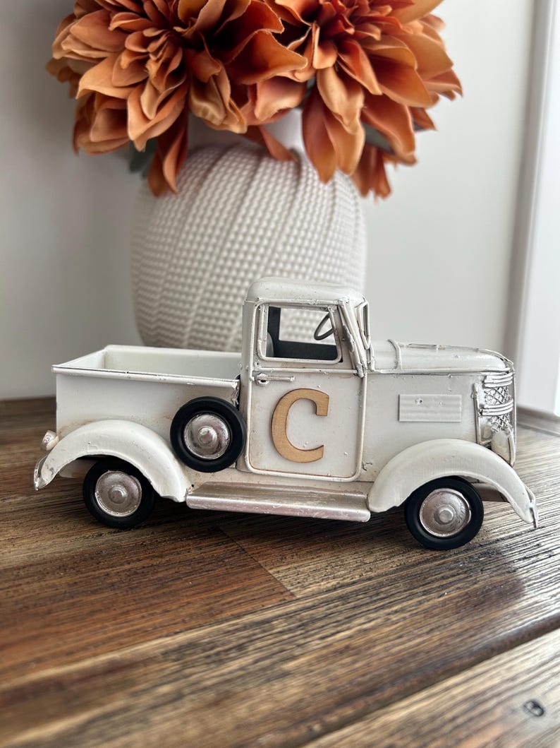 Peut inclure: Un camion en m&eacute;tal blanc de style vintage avec des roues noires et un "C" dor&eacute; sur la porte. Le camion est un objet d&eacute;coratif, peut-&ecirc;tre pour la d&eacute;coration int&eacute;rieure ou les expositions saisonni&egrave;res. Le camion est pos&eacute; sur une surface en bois.