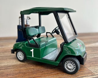 May include: A miniature dark green golf cart featuring a white number 1 on the side, a black roof, a clear windshield, and silver-toned wheels. The rear of the vehicle includes a dark blue golf bag containing silver-colored golf clubs.