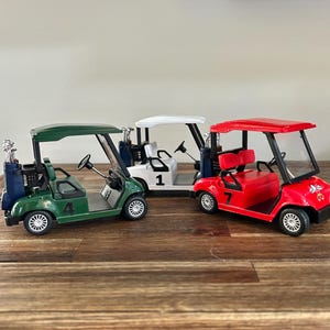 May include: Three miniature golf carts in green, white, and red, each featuring a steering wheel, seats, and a rear golf bag holder. The green cart is marked with the number 4, the white cart with the number 1, and the red cart with the number 7.