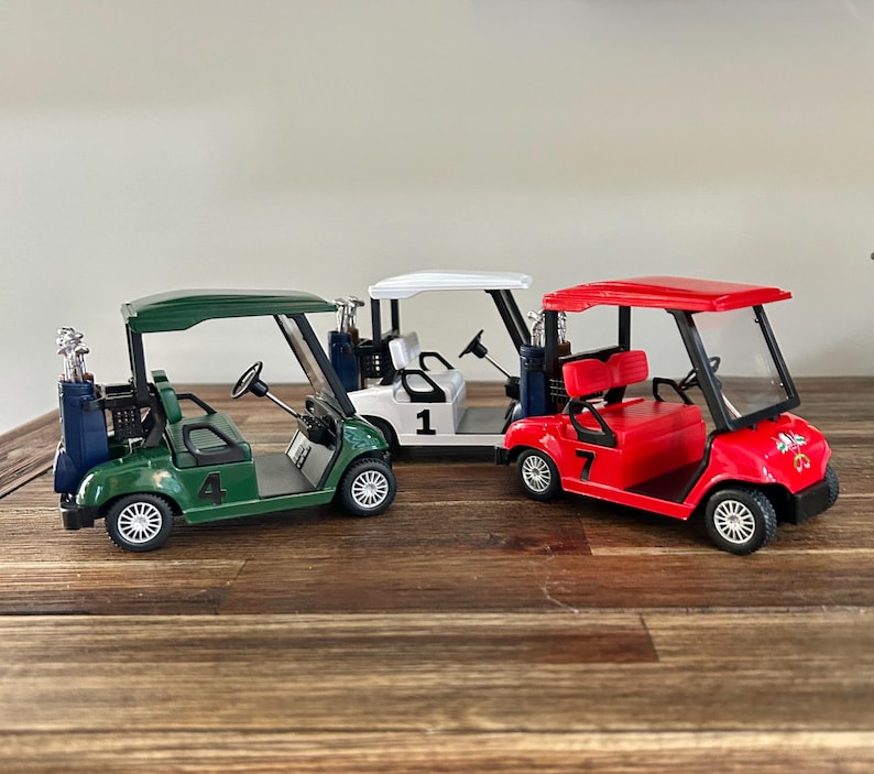 May include: Three miniature golf carts in green, white, and red, each featuring a steering wheel, seats, and a rear golf bag holder. The green cart is marked with the number 4, the white cart with the number 1, and the red cart with the number 7.